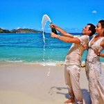 A couple on their honeymoon in Seychelles, standing on the beach and toasting with champagne, surrounded by serene ocean views.
